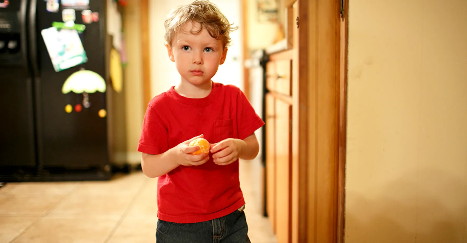 A little boy with a sad look on his face stands in his kitchen