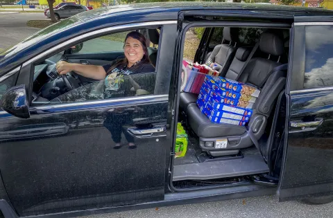 A woman smiles behind the steering wheel of a van loaded with food.